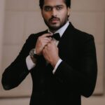 Portrait of a stylish man in a black tuxedo adjusting his bow tie indoors.
