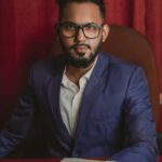 Confident man wearing a blue suit and eyeglasses sits at a desk with a serious expression.